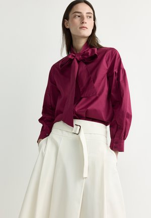 Woman wearing a dark pink blouse with a large bow tie and white high-waisted pleated skirt with belt, hands in pockets, looking sideways.