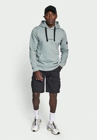 Homem a usar um hoodie cinza claro, boné preto, calções de carga pretos, meias brancas e sapatilhas brancas, de pé contra um fundo simples.