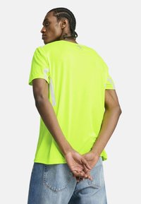 Neon żółta koszulka z krótkim rękawem z odblaskowymi akcentami na rękawach; gładka tekstura i luźny krój. Noszona z niebieskimi dżinsami.