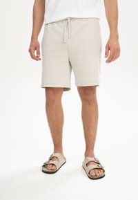 Beige katoenen shorts met elastische tailleband en koord, gekenmerkt door een ontspannen pasvorm en zijzakken, gecombineerd met beige sandalen.
