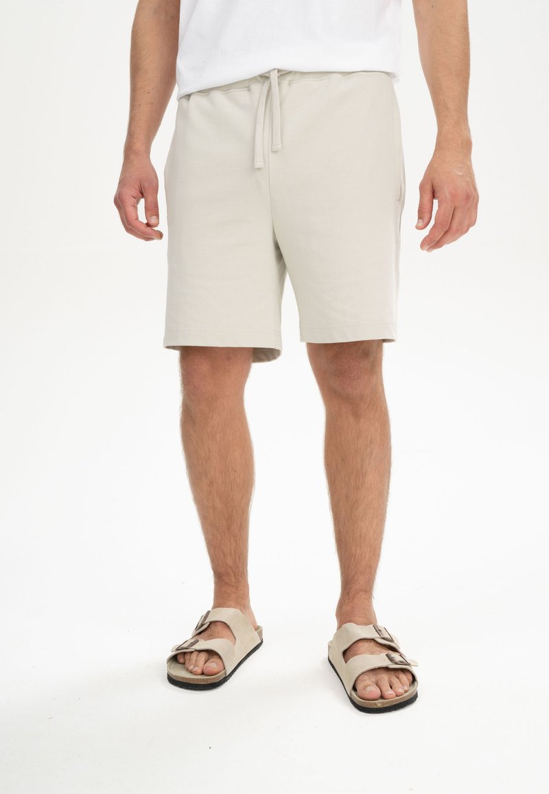 Beige katoenen shorts met elastische tailleband en koord, gekenmerkt door een ontspannen pasvorm en zijzakken, gecombineerd met beige sandalen.