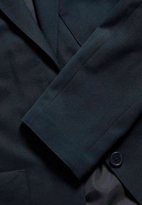 Tmavě modrý blazer s texturovanou tkaninou. Vybaven klopou s výstřihem, zapínáním na jeden knoflík a viditelnými detaily kapes.
