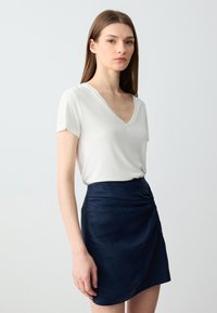 Weißes V-Ausschnitt-T-Shirt mit kurzen Ärmeln, kombiniert mit einem dunkelblauen Wickelrock, der vorne mit einem Knotendetail versehen ist und einen geschichteten Look erzeugt.