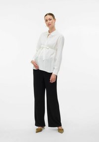 Chemise blanche à manches longues avec un nœud au niveau de la taille, associée à un pantalon large noir et des talons vert olive. Tissu lisse, coupe décontractée.