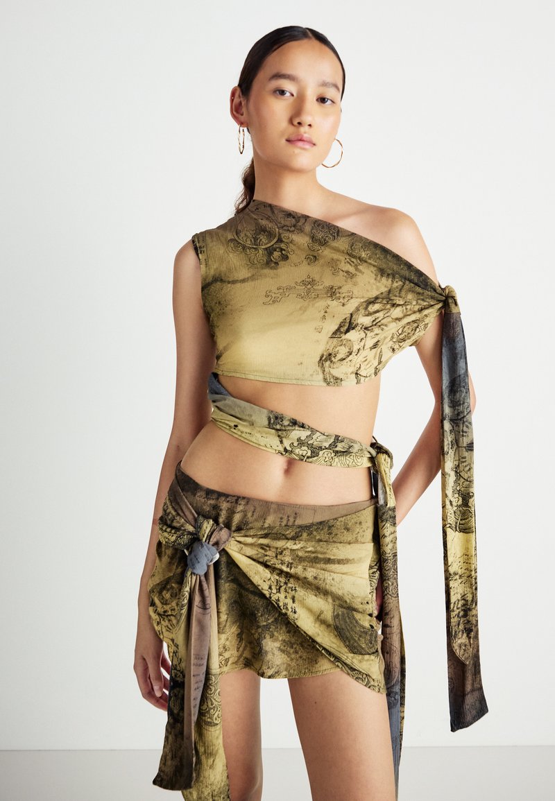 Junge Frau, die ein asymmetrisches Crop Top mit abstraktem Muster und einen passenden Wickelrock mit gebundenen Details trägt, steht vor einem weißen Hintergrund.