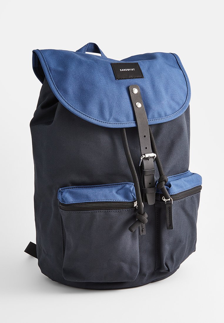 Rugzak met een blauwe flap en een donkerblauwe body, vervaardigd uit stevig materiaal. Bevat twee ritszakken aan de voorkant en een leren banddetail.