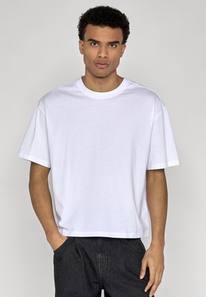 Ung mand med krøllet hår iført en ensfarvet hvid, stor T-shirt og sorte bukser, stående mod en lys grå baggrund.