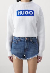 Maglione bianco a costine con logo "HUGO" in blu sul petto, abbinato a pantaloni shorts in denim sbiadito con orli sfrangiati e tasche.