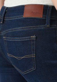 Tmavomodré džínsy z denimu s koženou nášivkou značky, zlatým prešívaním a klasickým dizajnom vreciek s jemným zakrivením na vrchu.