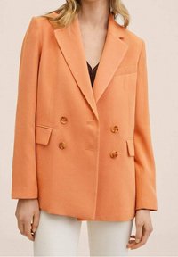 Femme portant un blazer orange boutonné double avec des boutons marron, par-dessus un haut sombre et un pantalon blanc, se tenant devant un fond neutre.