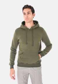 Uomo che indossa un felpa verde olivo con tasca frontale e cordini, abbinata a pantaloni cargo grigio chiaro, in piedi davanti a uno sfondo bianco.