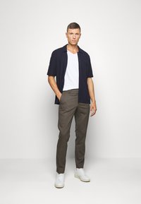 Model i en mørk kortærmet skjorte over en hvid V-hals t-shirt, parret med brune ternede bukser og hvide sneakers. Enkel, ren design.