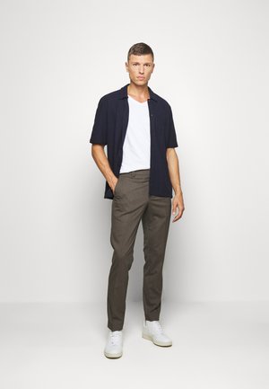 Modèle portant une chemise à manches courtes noire sur un t-shirt blanc à col en V, associé à un pantalon à carreaux marron et des baskets blanches. Design simple et épuré.