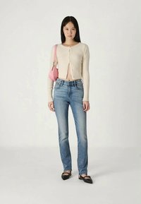 Cardigã curto em bege com fecho de botão, combinado com jeans azul claro de perna reta. Os acessórios incluem uma pequena mala rosa e sapatilhas pretas.