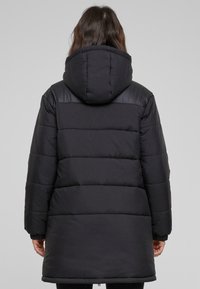 Abrigo de plumón negro con capucha, con secciones cosidas horizontalmente, puños elásticos y un diseño de longitud media. Acabado mate y suave.