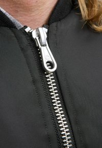 Veste en tissu noir avec une fermeture éclair argentée visible. La fermeture éclair est dotée d'une tirette lisse et de dents en métal le long de l'ouverture avant, garantissant sa durabilité.