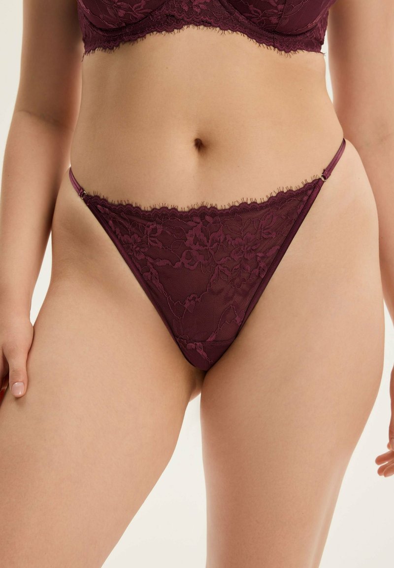 Mauve Spitzenstring mit floralem Muster, transparenten Netzpaneelen, zarten Fransen an den Kanten und minimalistischen Trägern mit einem kleinen metallenem Akzent.
