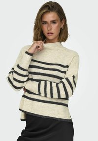Pull rayé crème et noir avec un col montant, texture tricotée et manches longues, présentant un dos légèrement plus long.