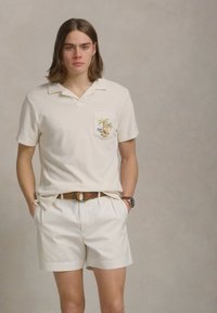 Bela polo majica s žepom na prsih, okrašena z vezenjem palma, v kombinaciji s smetanastimi kratkimi hlačami in rjavim pasom. Preprost, sproščen dizajn.