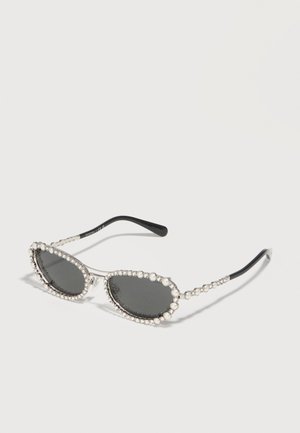 Lunettes de soleil ovales noires avec montures argentées ornées de perles et de petits strass le long des bords et des branches, présentées sur une surface blanche.