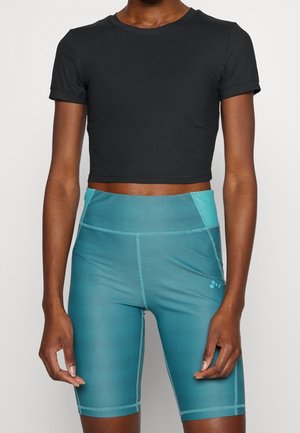 Persona que lleva una camiseta corta negra de manga corta y unos pantalones cortos de ciclismo texturizados turquesa de talle alto, de pie contra un fondo liso.