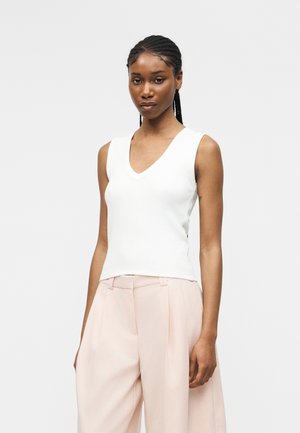 Femme aux cheveux tressés portant un haut blanc sans manches à col en V et un pantalon large rose clair, debout devant un fond uni.