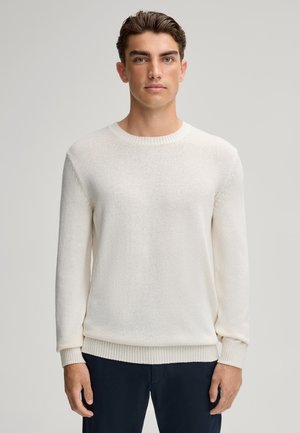 Weißer Strickpullover mit Rundhalsausschnitt, gerippten Bündchen und Säume, mit einer strukturierten Oberfläche und einem schlichten Design. Getragen über dunklen Hosen.