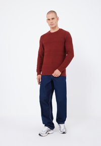 Czerwony sweter z teksturowanym dzianinowym splotem, z okrągłym dekoltem, w połączeniu z ciemnoniebieskimi, luźnymi dżinsami oraz srebrnymi butami sportowymi. Model stoi na tle białej ściany.