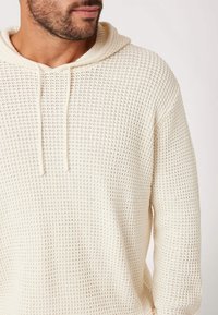 Crème gebreide hoodie met een textuurpatroon, koordjes en een ontspannen pasvorm, die een ademende geweven stof en ribbelranden toont.