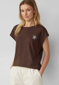 Woman with long blonde hair wearing a brown short-sleeve shirt with a small turtle design and light-colored pants with hands in pockets.