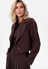 Burgundowa marynarka ze streczowym wzorem w paski, skrócona, z usztywnionym kołnierzem, z zapięciem na jeden guzik, w połączeniu z czarną bluzką w kwiaty.