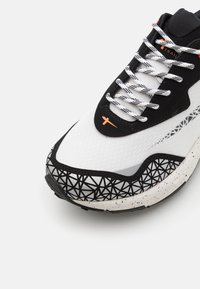 Zapatilla atlética blanca y negra con un patrón geométrico negro, parte superior blanca texturizada, detalles en ante negro y cordones reflectantes.