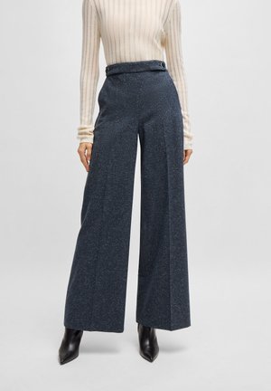 Weite Hosen aus dunkelblauem, gepunktetem Stoff mit hoher Taille und Akzentknöpfen. Kombiniert mit einem hellen, gerippten Langarmoberteil.