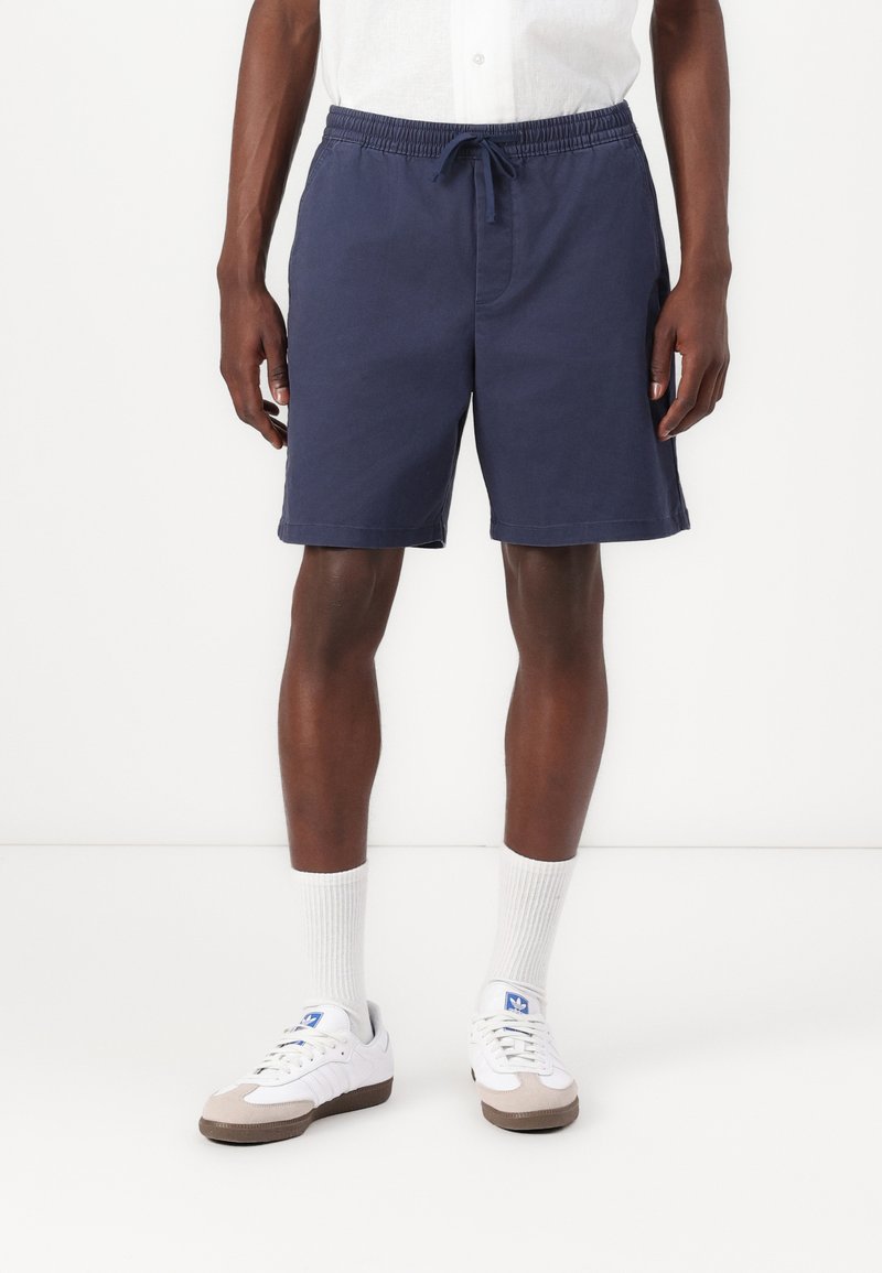Marineblå shorts med elastisk midje og snor. Lomme på siden, avslappet passform. Brukt med hvite sokker og joggesko.