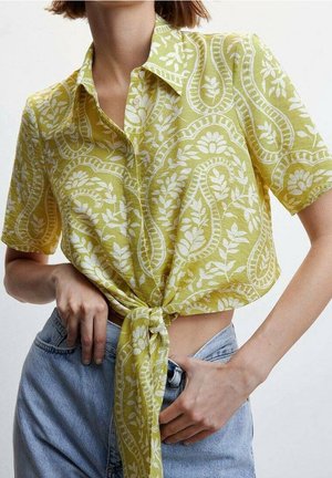 Personne portant une chemise à manches courtes vert citron avec des motifs floraux cachemire blancs, nouée à la taille, et un jean bleu clair.
