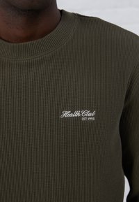 Suéter de canalé verde oliva con cuello redondo, que presenta texto bordado en blanco "Health Club EST 1998" en la parte izquierda del pecho.