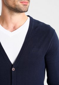 Mann in dunkelblauem, geknöpftem Cardigan über einem weißen V-Ausschnitt-T-Shirt, zeigt Brust und Untergesicht, schaut zur Seite.
