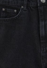 Gros plan sur un jean en denim noir montrant une poche avant avec des rivets en métal, un passant de ceinture et une ceinture avec un bouton métallique visible.