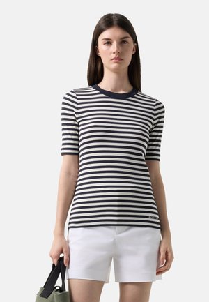 Vrouw met een zwart-wit gestreept T-shirt met korte mouwen en witte korte broek, die een groene en zwarte handtas vasthoudt tegen een effen achtergrond.