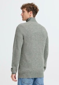 Maglione grigio a costine con collo alto, maniche lunghe e tessuto strutturato, visto da dietro, abbinato a jeans in denim blu.