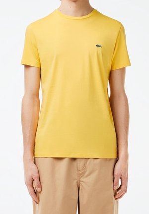 Gul bomulds t-shirt med rund hals og korte ærmer. Har et lille grønt geometrisk logo på venstre bryst. Glat tekstur.