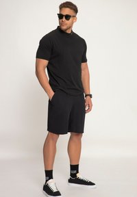 Camiseta negra de manga corta y pantalones cortos negros con bolsillos. Zapatillas negras con cordones blancos y calcetines con estampado. Gafas de sol completan el look.