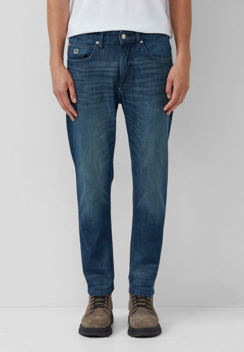 Dark blue denim jeans with a straight cut, featuring whisker patterns, five-pocket design, and a metal button closure. Worn with brown boots.