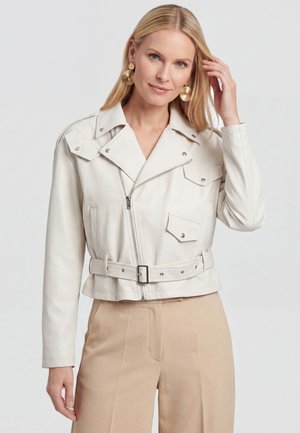 Blonde Frau mit goldfarbenen, runden Ohrringen, einer weißen, gürtelförmigen Lederjacke und beigen, hoch taillierten Hosen, die mit einer Hand ihr Haar berührt.