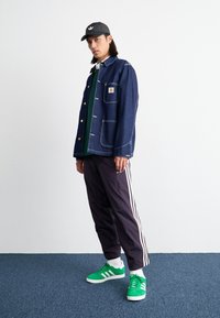 Denim jacket with gold buttons and front pockets, green shirt underneath, dark sweatpants with white stripes, and bright green sneakers.