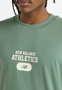 Grøn bomulds T-shirt med teksten "NEW BALANCE ATHLETICS" i hvid, med et rundt logo nedenfor. Blød tekstur og klassisk crew neck design.