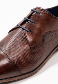 Zapato de cuero marrón con una superficie texturizada, cordones azules y un logo sutil. Presenta una punta redondeada y detalles en contraste.