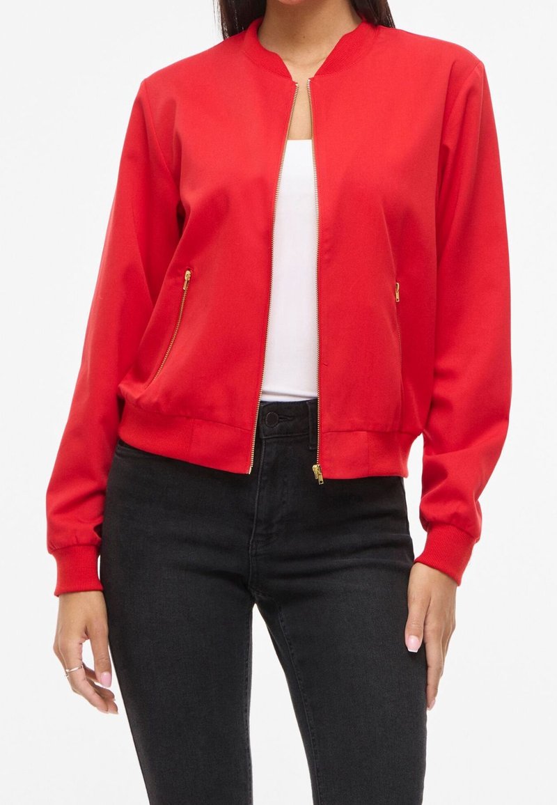 Femme portant une veste bomber rouge vif zippée avec des fermetures éclair dorées, un top blanc en dessous et un jean noir taille haute.