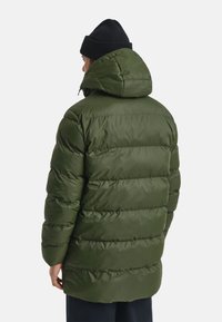Man in een olijfgroene gewatteerde winterjas met capuchon en een zwarte gebreide beanie, gezien vanachter tegen een effen achtergrond.