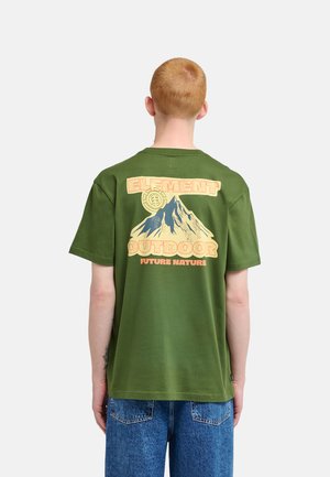 Personne aux cheveux rouges courts portant un t-shirt vert avec un motif de montagne et les mots "Element Outdoor Future Nature" dans le dos.